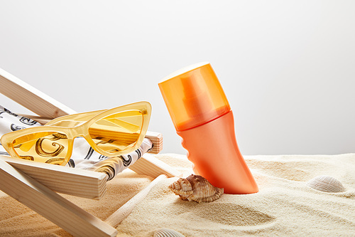 orange sunscreen in sand near seashells, yellow sunglasses and deck chair on grey background