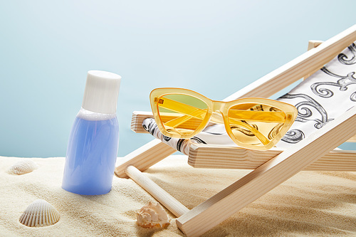 blue lotion in sand near seashells, yellow sunglasses and deck chair on blue background