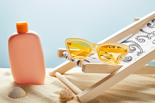 sunscreen lotion in sand near seashells, yellow sunglasses and deck chair on blue background
