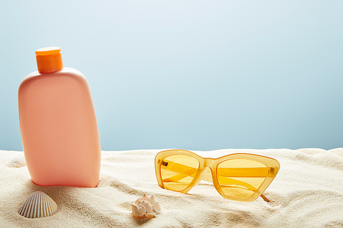 sunscreen lotion in sand near seashells and yellow sunglasses on blue background