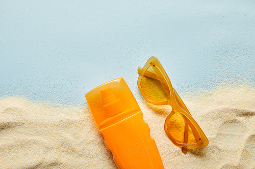 top view of sunscreen in orange bottle near sunglasses on blue background with sand