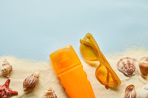 top view of sunscreen in orange bottle near sunglasses on blue background with sand and seashells