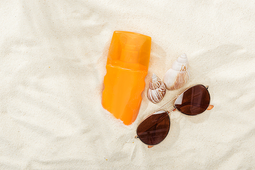 orange bottle of sunscreen on sand with seashells and stylish sunglasses