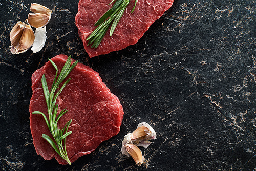 top view of uncooked beef sirloins with garlic and rosemary on black marble background