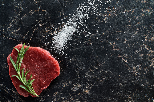 top view of raw beef steak with rosemary twig on black marble surface with scattered salt crystals