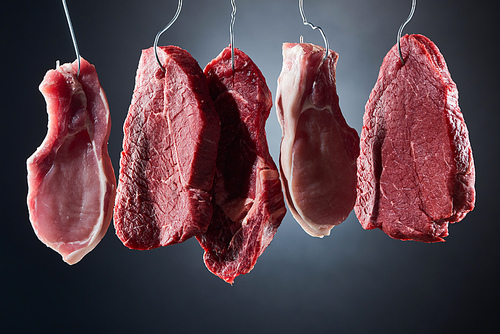 assorted raw meat steaks on metal hooks on dark black background