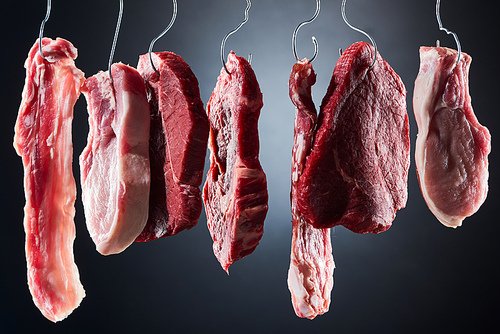 assorted raw meat and bacon on metal hooks on dark black background