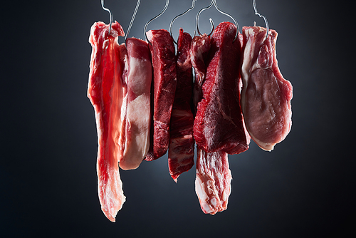 assorted raw meat steaks and bacon on metal hooks on dark black background
