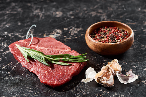 raw beef steak with rosemary twig near small bowl with peppercorns mix and garlic on black marble surface