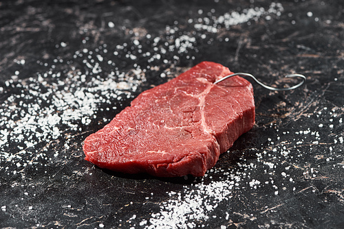 raw meat steak with metal hook on black marble surface with scattered salt