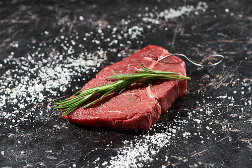 raw meat steak with rosemary twig on black marble surface with scattered salt