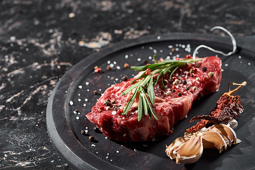 raw meat steak with rosemary twig sprinkled with salt and pepper near garlic and dried cayenne on round wooden surface