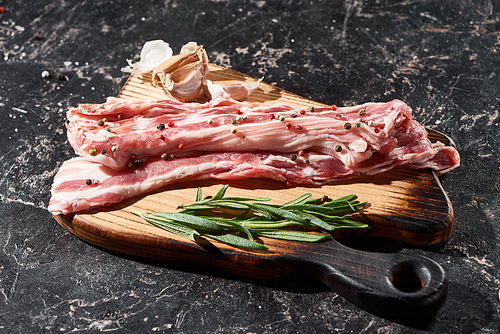wooden cutting board with raw pork slices near rosemary and garlic on black marble surface