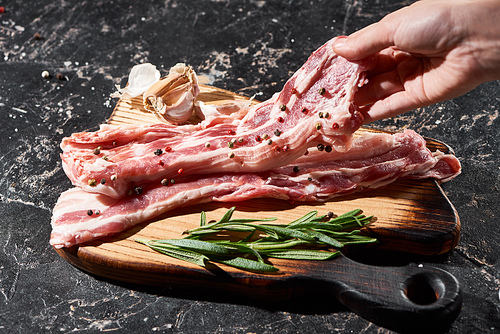 cropped view of raw pork slice in hand near rosemary and garlic on black marble surface