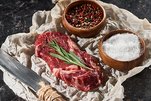 raw beef steak near knife and wooden bowls with salt and pepper on black marble surface