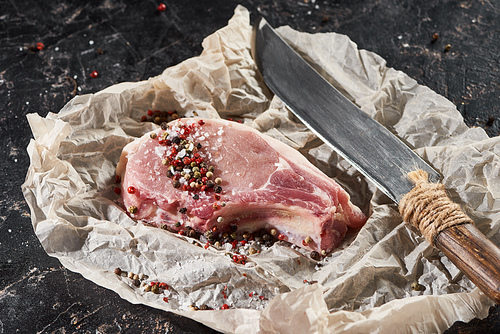 raw pork steak sprinkled with salt and pepper near knife on parchment paper on black marble surface