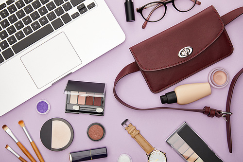 top view of bag and decorative cosmetics near laptop isolated on violet