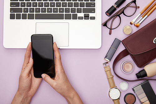 top view of woman holding smartphone with blank screen near laptop and decorative cosmetics isolated on violet