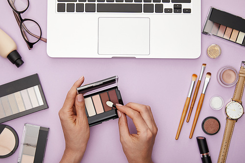 cropped view of woman holding eye shadow near laptop and decorative cosmetics isolated on violet