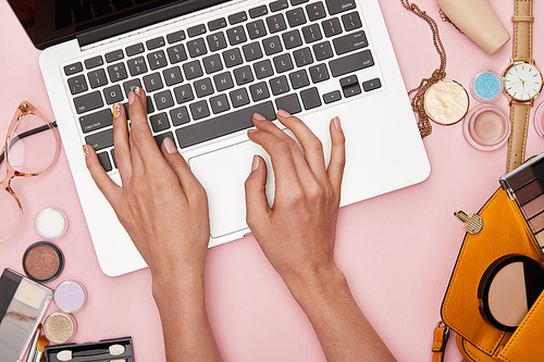 top view of woman typing on laptop near decorative cosmetics and necklace isolated on pink