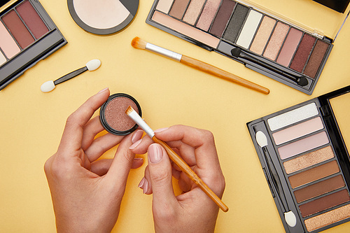 top view of woman holding cosmetic brush near eye shadow isolated on yellow