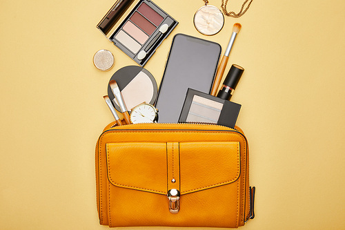 top view of bag with smartphone and decorative cosmetics isolated on yellow