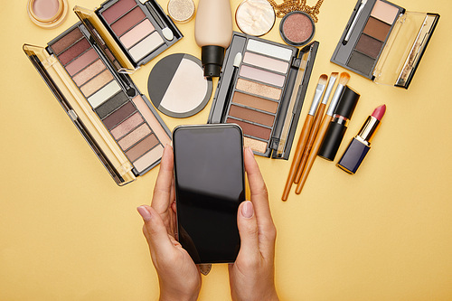 cropped view of woman holding smartphone with blank screen near decorative cosmetics isolated on yellow