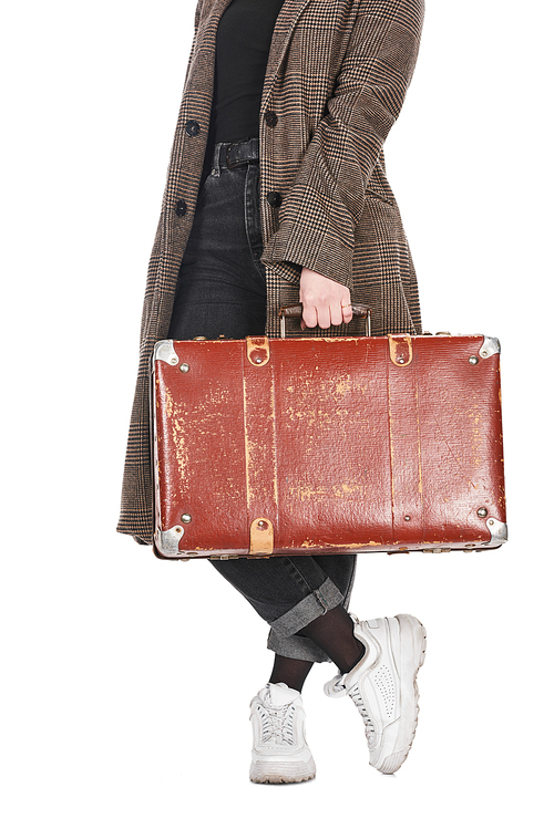 cropped view of woman in plaid coat with crossed legs holding vintage weathered suitcase isolated on white
