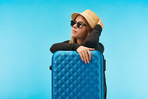 blonde girl in sunglasses and straw hat near blue suitcase isolated on blue