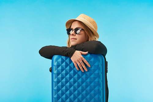 dreamy blonde girl in sunglasses and straw hat near blue travel bag isolated on blue