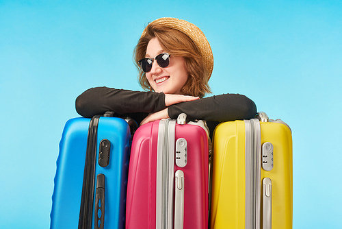 happy girl in sunglasses and straw hat near multicolored travel bags isolated on blue