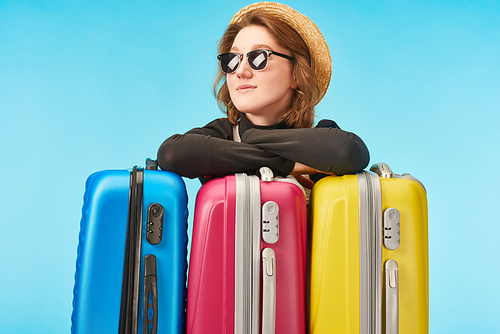 smiling girl in sunglasses and straw hat near multicolored travel bags isolated on blue