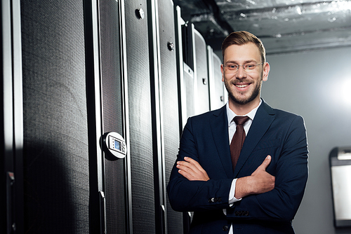 selective focus of cheerful businessman in suit and glasses standing with crossed arms in data center