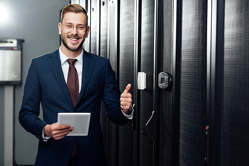 selective focus of cheerful businessman holding digital tablet and showing thumb up in data center