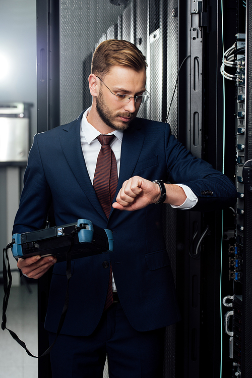 handsome businessman standing with reflectometer and looking at watch in server room