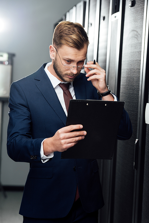 handsome businessman touching glasses while looking at clipboard