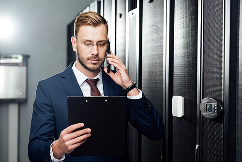handsome businessman talking on smartphone while looking at clipboard in server room