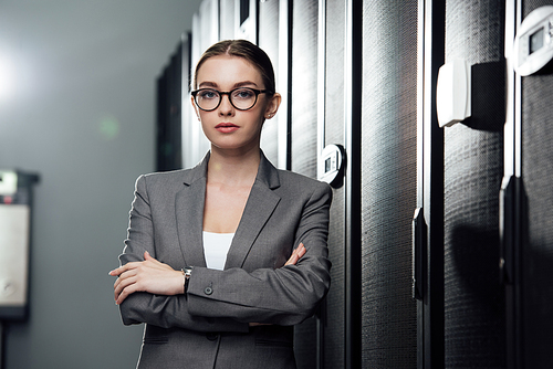 selective focus of attractive businesswoman in glasses standing with crossed arms in data center