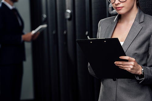 cropped view of businesswoman holding clipboard near businessman and server racks