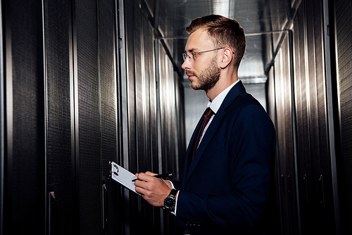 side view of businessman in glasses holding clipboard near server racks