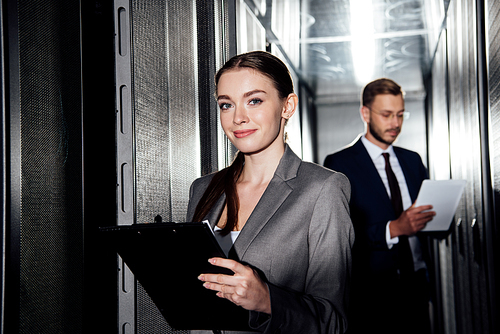 selective focus of cheerful businesswoman holding clipboard near bearded businessman in data canter