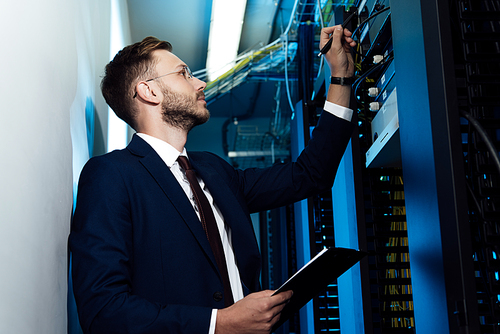 side view of bearded businessman in glasses holding pen and clipboard while looking at server rack