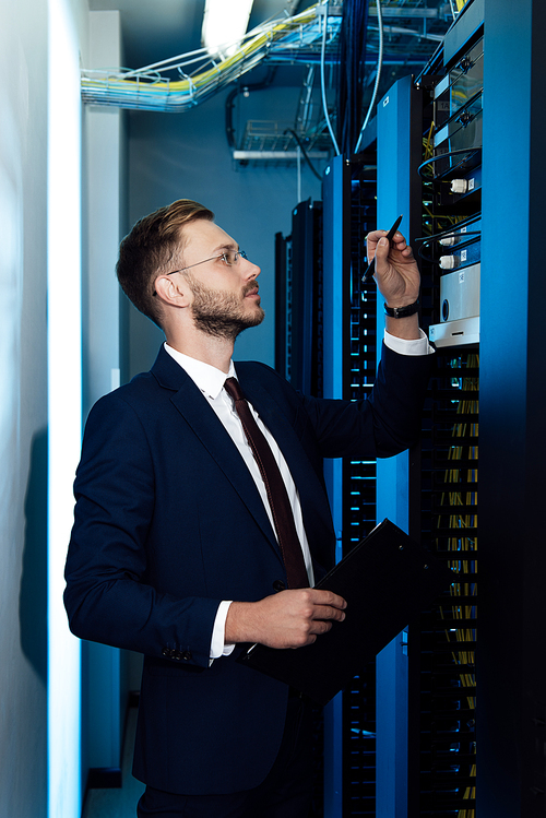 profile of handsome businessman in glasses holding pen and clipboard while looking at server rack