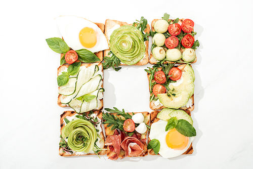 Top view of toasts with cut vegetables, fried eggs and prosciutto on white surface