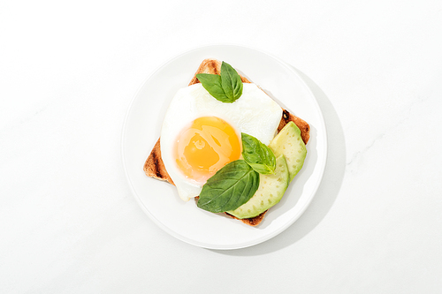 Top view of toast with fried egg and cut avocado on plate on white surface