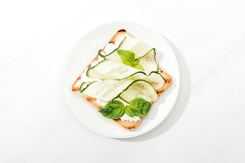 Top view of toast with sliced cucumber and basil on plate on white surface