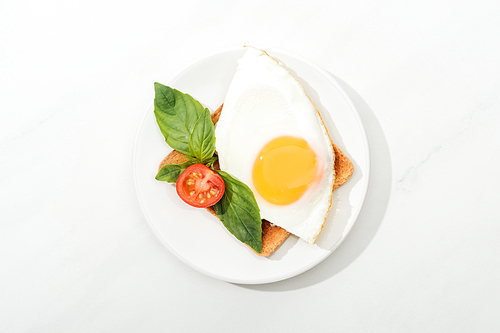 Top view of toast with fried egg, basil and cut tomato cherry on plate on white surface