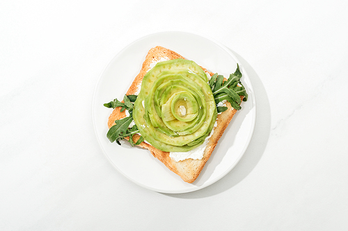 Top view of toast with sliced avocado and arugula on plate on white surface