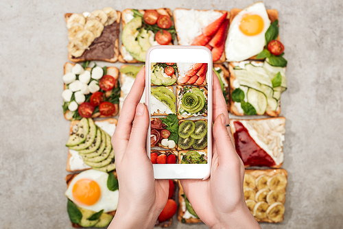 Partial view of woman holding smartphone and taking photo of toasts with fruits and vegetables