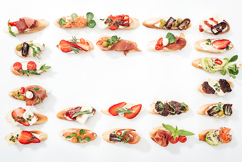 top view of frame made of traditional italian bruschetta with prosciutto, salmon, fruits, s and herbs on white